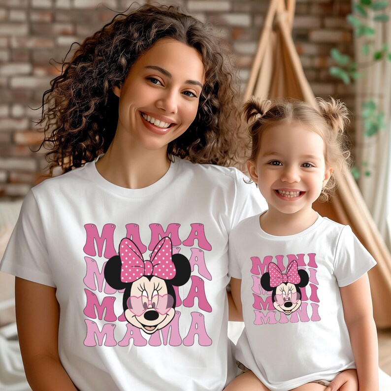 May include: A white t-shirt with a pink Minnie Mouse graphic and the text "MAMA" repeated multiple times. A smaller white t-shirt with a pink Minnie Mouse graphic and the text "MINI" repeated multiple times. The shirts are worn by a woman and a young girl.
