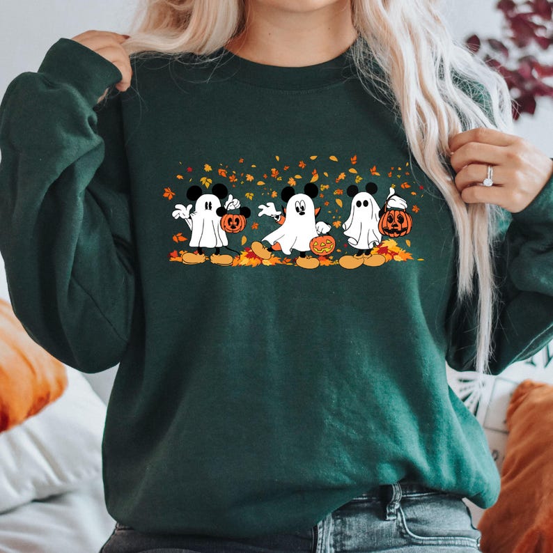 May include: A green sweatshirt with a graphic of three Mickey Mouse ghosts holding jack-o'-lanterns and walking through a field of autumn leaves.
