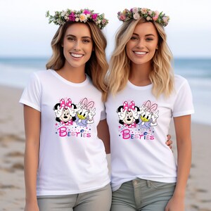 May include: Two women wearing white t-shirts with a graphic of Minnie Mouse and Daisy Duck. The shirts say "Besties" in pink letters.
