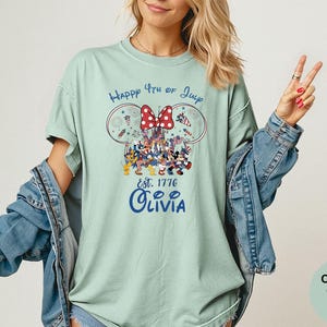 Pode incluir: Camiseta verde claro com um gr&aacute;fico do 4 de julho. O design inclui orelhas do Mickey e da Minnie, um la&ccedil;o vermelho e o texto "Happy 4th of July Est. 1776 Olivia". A camisa &eacute; Comfort Colors Bay.