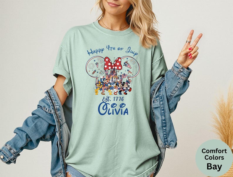 Pode incluir: Camiseta verde claro com um gr&aacute;fico do 4 de julho. O design inclui orelhas do Mickey e da Minnie, um la&ccedil;o vermelho e o texto "Happy 4th of July Est. 1776 Olivia". A camisa &eacute; Comfort Colors Bay.