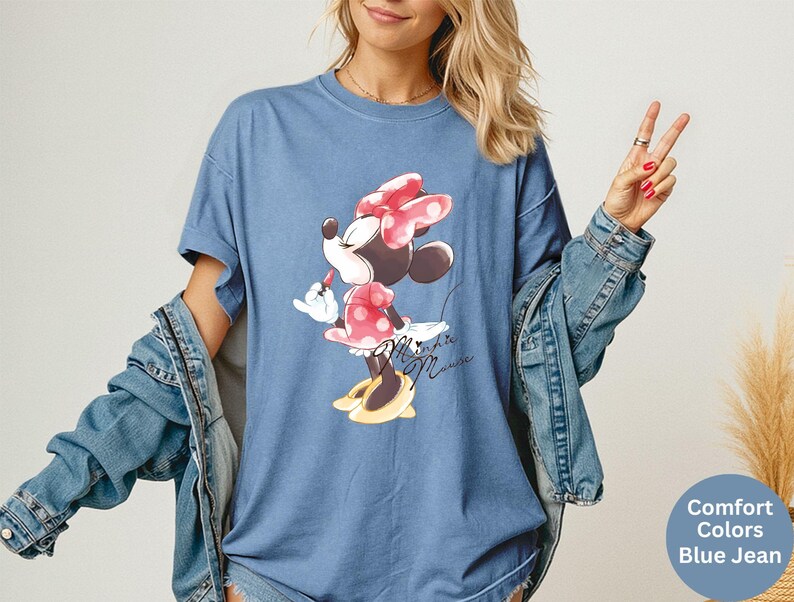 K&ouml;nnte beinhalten: Blaues Jeans-T-Shirt mit Minnie Mouse-Grafik. Die Zeichentrickfigur wird beim Auftragen von Lippenstift gezeigt, tr&auml;gt ein gepunktetes Kleid und eine Schleife. Die Worte "Minnie Mouse" sind darunter in Kursivschrift geschrieben.