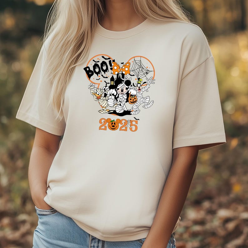 May include: Beige t-shirt with a Halloween graphic. The design features Mickey and Minnie Mouse as mummies, plus other characters, the word "Boo!", a spider web, and the year "2025" in orange.