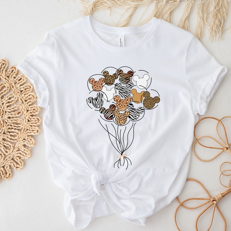 Puede incluir: Camiseta blanca con un gr&aacute;fico de un mont&oacute;n de cabezas de Mickey Mouse con estampado de animales en forma de globos.