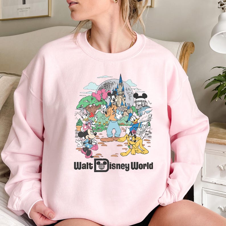 May include: Pink sweatshirt with a graphic print of Mickey Mouse and other Disney characters in front of Cinderella's Castle. The text "Walt Disney World" is printed below the graphic.