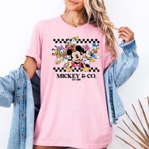 May include: Pink t-shirt featuring a graphic of Mickey Mouse and friends, including Donald Duck, Goofy, and Minnie Mouse, with the text "Mickey & Co. EST 1928" in black. The design is set against a black and white checkered pattern.