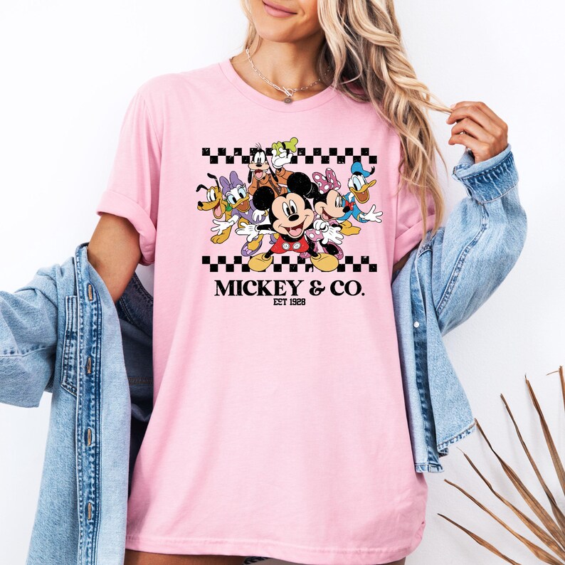 May include: Pink t-shirt featuring a graphic of Mickey Mouse and friends, including Donald Duck, Goofy, and Minnie Mouse, with the text "Mickey & Co. EST 1928" in black. The design is set against a black and white checkered pattern.