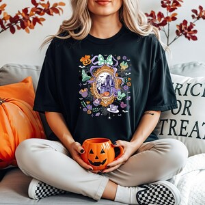 Puede incluir: Camiseta negra con un dise&ntilde;o de Halloween que presenta un castillo, calabazas y otros elementos festivos. La camiseta tiene un dise&ntilde;o de espejo decorativo y personajes de dibujos animados. Una persona sostiene una taza naranja con forma de calabaza.