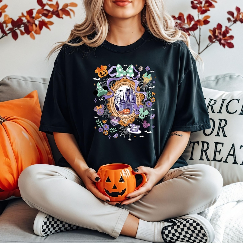 Puede incluir: Camiseta negra con un dise&ntilde;o de Halloween que presenta un castillo, calabazas y otros elementos festivos. La camiseta tiene un dise&ntilde;o de espejo decorativo y personajes de dibujos animados. Una persona sostiene una taza naranja con forma de calabaza.