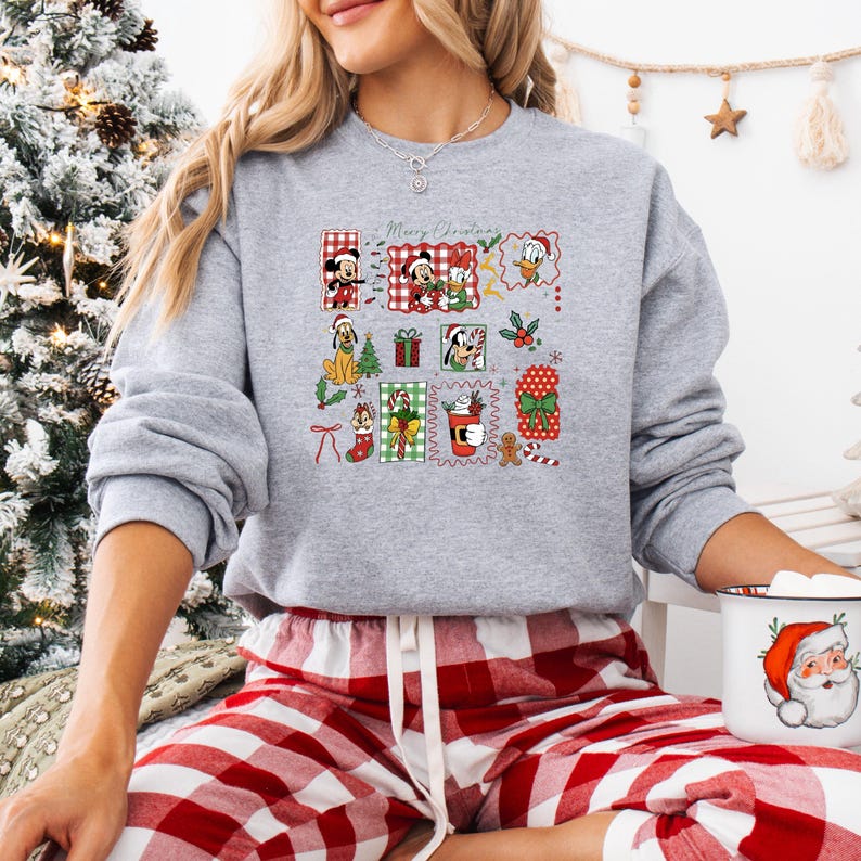 May include: A gray sweatshirt with a colorful Christmas-themed graphic featuring cartoon characters and the text "Merry Christmas." The design includes images of Mickey and Minnie Mouse, Donald Duck, and other festive elements. The person is wearing red and white plaid pajama pants.