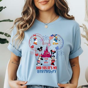 May include: Light blue t-shirt featuring a colorful graphic of Disney characters celebrating the 4th of July and a birthday. The design includes Mickey and Minnie Mouse, Donald and Daisy Duck, a castle, and the text "AND YES IT'S MY BIRTHDAY".