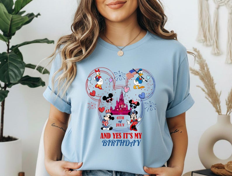 May include: Light blue t-shirt featuring a colorful graphic of Disney characters celebrating the 4th of July and a birthday. The design includes Mickey and Minnie Mouse, Donald and Daisy Duck, a castle, and the text "AND YES IT'S MY BIRTHDAY".