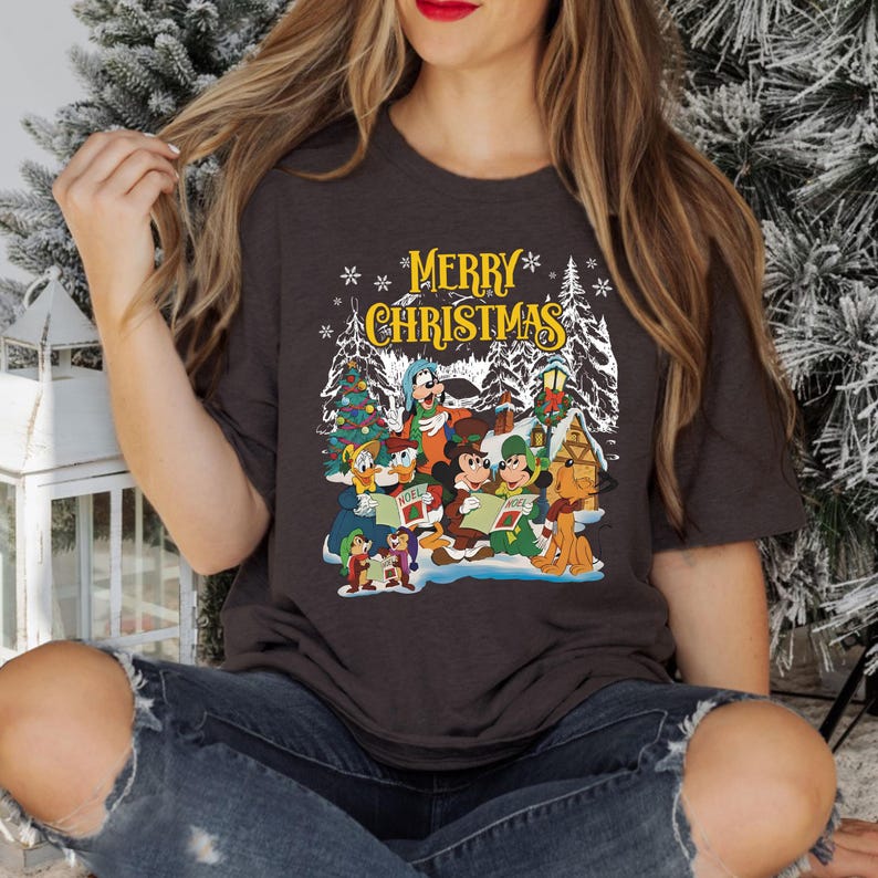 May include: A dark gray t-shirt with a Christmas-themed design featuring Mickey Mouse, Donald Duck, Goofy, Pluto, and other Disney characters singing carols. The text "Merry Christmas" is printed in yellow above the characters.
