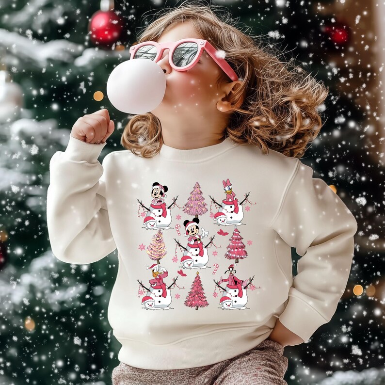 May include: A cream-coloured sweatshirt with a festive design of Disney characters as snowmen, with pink Christmas trees. The child in the image is wearing pink sunglasses and blowing a bubble. The background is a snowy Christmas scene.