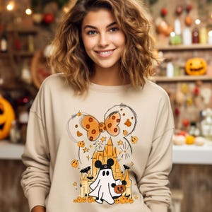May include: A beige sweatshirt with a Halloween-themed graphic featuring a ghost wearing Mickey Mouse ears, a pumpkin, and a castle. The graphic also includes a large orange and brown bow with white polka dots.