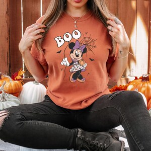 May include: Orange t-shirt with a cartoon image of Minnie Mouse dressed in a purple and black Halloween costume. The shirt says "BOO" in large white letters.