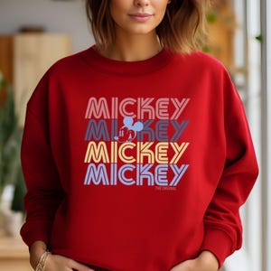May include: A red sweatshirt featuring the word "MICKEY" stacked four times in different colors, with a Mickey Mouse graphic. The text "THE ORIGINAL" is below the stacked text. The sweatshirt is a crew neck style.