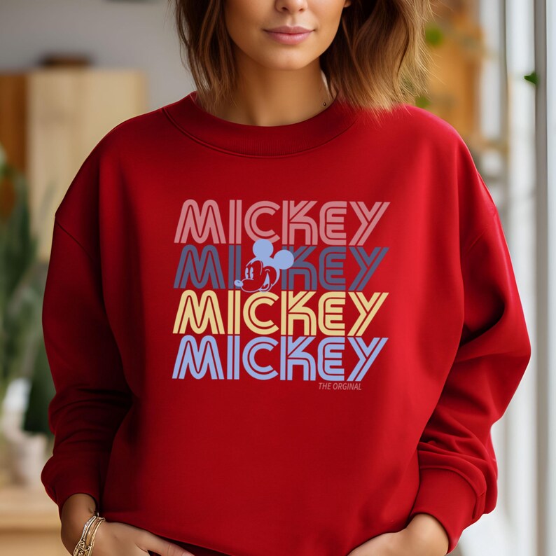 May include: A red sweatshirt featuring the word "MICKEY" stacked four times in different colors, with a Mickey Mouse graphic. The text "THE ORIGINAL" is below the stacked text. The sweatshirt is a crew neck style.