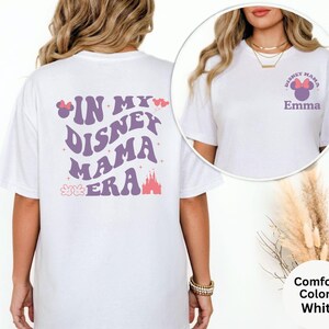 Puede incluir: Camiseta blanca con el texto "IN MY DISNEY MAMA ERA" en morado, rosa y rojo. La parte delantera de la camiseta tiene el texto "DISNEY MAMA" con un lazo de Minnie Mouse y el nombre "Emma". La camiseta es de la marca Comfort Colors.