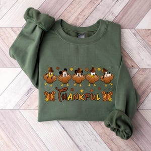 May include: Green sweatshirt featuring a Thanksgiving design. The design includes cartoon turkey characters wearing pilgrim hats, with the word "THANKFUL" in brown script. Autumn leaves and pumpkins are also included in the design.