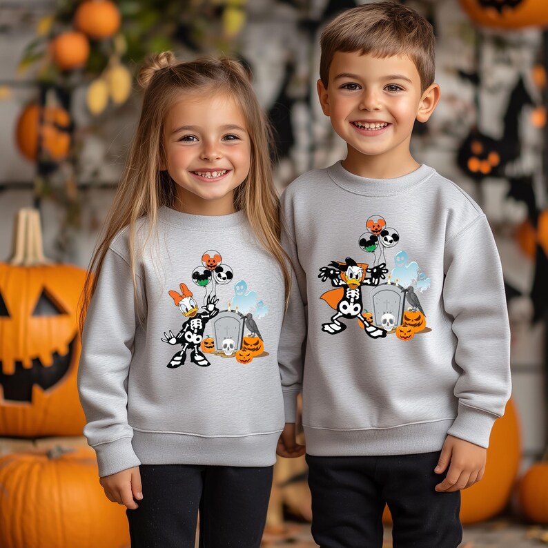 May include: Two children wearing light gray Halloween sweatshirts. The sweatshirts feature a cartoon character dressed as a skeleton with balloons, pumpkins, and a tombstone. The background includes pumpkins and fall foliage.
