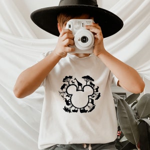 May include: White t-shirt featuring a black silhouette of Mickey Mouse ears formed by animals and trees. The text "Instax" is visible on a white instant camera held by the person wearing the shirt.
