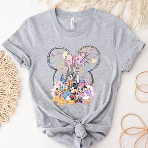 May include: Gray t-shirt with a colorful graphic print of Mickey Mouse, Minnie Mouse, Donald Duck, Goofy, and Pluto in front of Cinderella's Castle. The graphic is surrounded by a black outline of Mickey Mouse's ears with a pink bow.