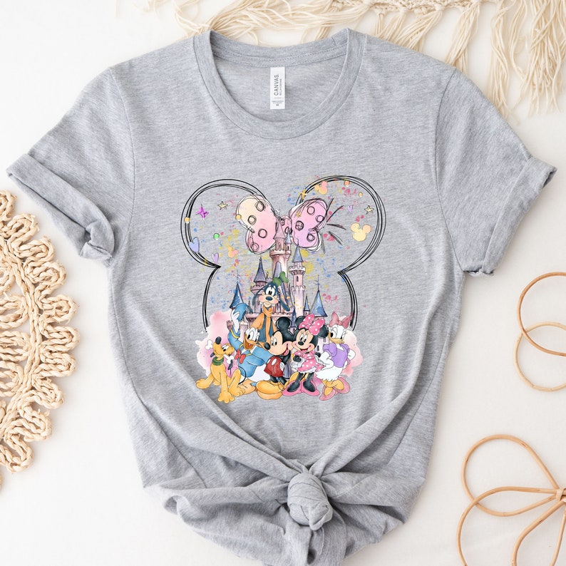 May include: Gray t-shirt with a colorful graphic print of Mickey Mouse, Minnie Mouse, Donald Duck, Goofy, and Pluto in front of Cinderella's Castle. The graphic is surrounded by a black outline of Mickey Mouse's ears with a pink bow.