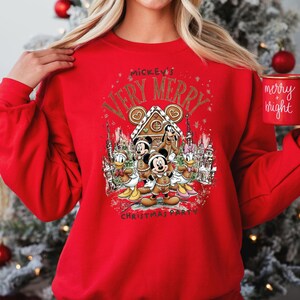 Puede incluir: Una sudadera roja con un dise&ntilde;o festivo que presenta a Mickey Mouse y sus amigos. El dise&ntilde;o incluye el texto "Mickey's Very Merry Christmas Party" y una casa de jengibre. Tambi&eacute;n se ve una taza roja con "merry + bright".