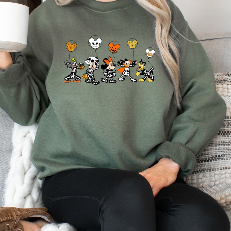 Op de afbeelding: Groen sweatshirt met een afbeelding van Mickey Mouse en zijn vrienden verkleed als skeletten die ballonnen vasthouden. De ballonnen zijn oranje, geel en wit met Mickey Mouse-oren.