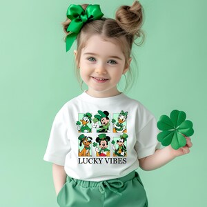 May include: A white t-shirt with a green shamrock design featuring Mickey Mouse, Minnie Mouse, Donald Duck, Goofy, and Pluto. The shirt has the text "LUCKY VIBES" printed on the front.