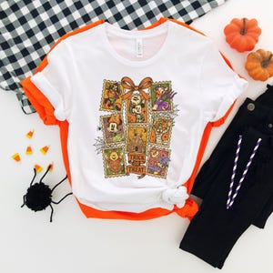 May include: White t-shirt with a Halloween-themed graphic featuring cartoon characters on postage stamp designs. The shirt has a round neck and short sleeves. Black pants, pumpkins, and candy corn are also in the image.
