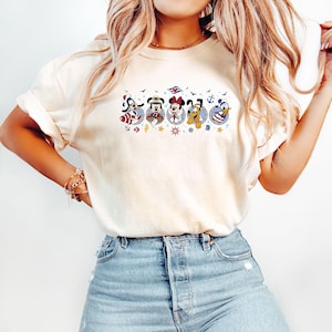 May include: Cream-colored t-shirt featuring a nautical-themed graphic of Disney characters, including Mickey Mouse, Minnie Mouse, and Donald Duck. The design incorporates anchors, stars, and a cruise ship logo. The shirt is paired with blue denim jeans.
