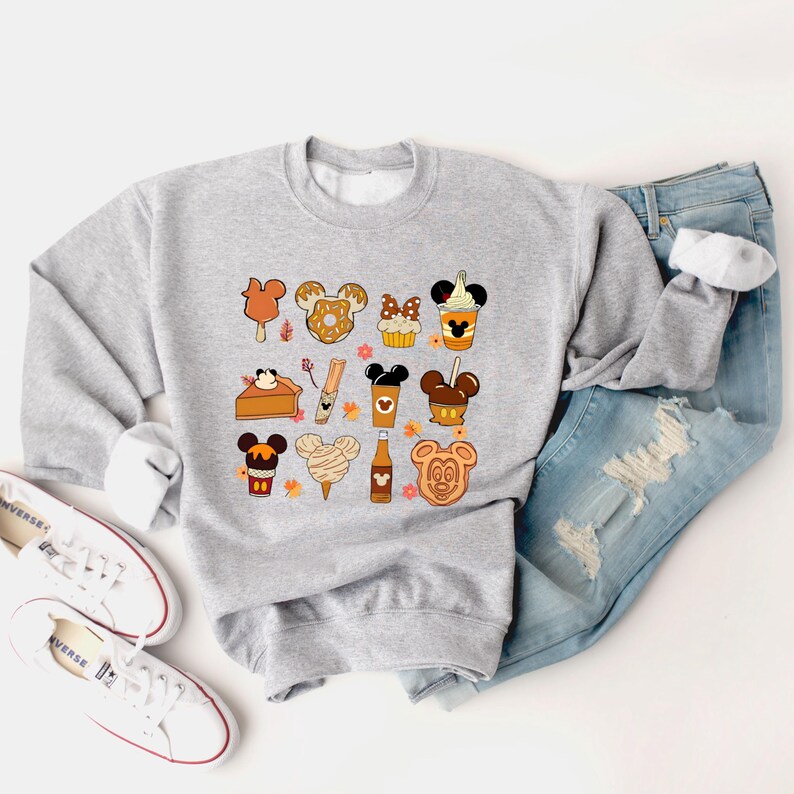 May include: A light gray sweatshirt featuring a colorful graphic of various fall-themed treats in the shape of a famous mouse. The design includes ice cream bars, donuts, cupcakes, and coffee cups. The sweatshirt is paired with blue jeans and white sneakers.