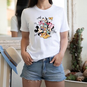 May include: White t-shirt featuring a colorful graphic of Disney characters including Mickey Mouse, Minnie Mouse, Donald Duck, Daisy Duck, Goofy, and Pluto. The shirt has character signatures and the text "Minnie Mouse 2020".