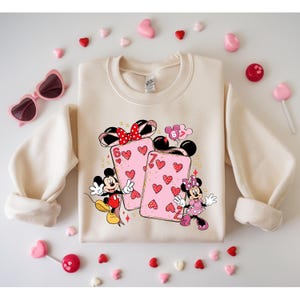 May include: Cream-colored sweatshirt featuring a graphic of Mickey and Minnie Mouse with playing cards. The cards are pink with heart designs. The sweatshirt is surrounded by heart-shaped candies, lollipops, and pink heart-shaped sunglasses.