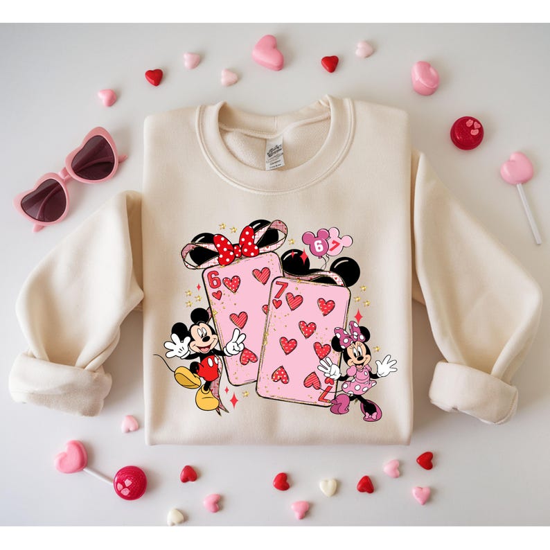 May include: Cream-colored sweatshirt featuring a graphic of Mickey and Minnie Mouse with playing cards. The cards are pink with heart designs. The sweatshirt is surrounded by heart-shaped candies, lollipops, and pink heart-shaped sunglasses.