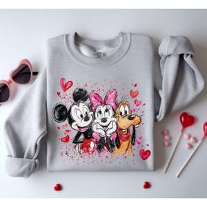 May include: A light gray sweatshirt featuring Mickey Mouse, Minnie Mouse, and Goofy, surrounded by red hearts and paint splatters. The cartoon characters are smiling and hugging. Pink heart-shaped sunglasses and heart-shaped lollipops are in the background.