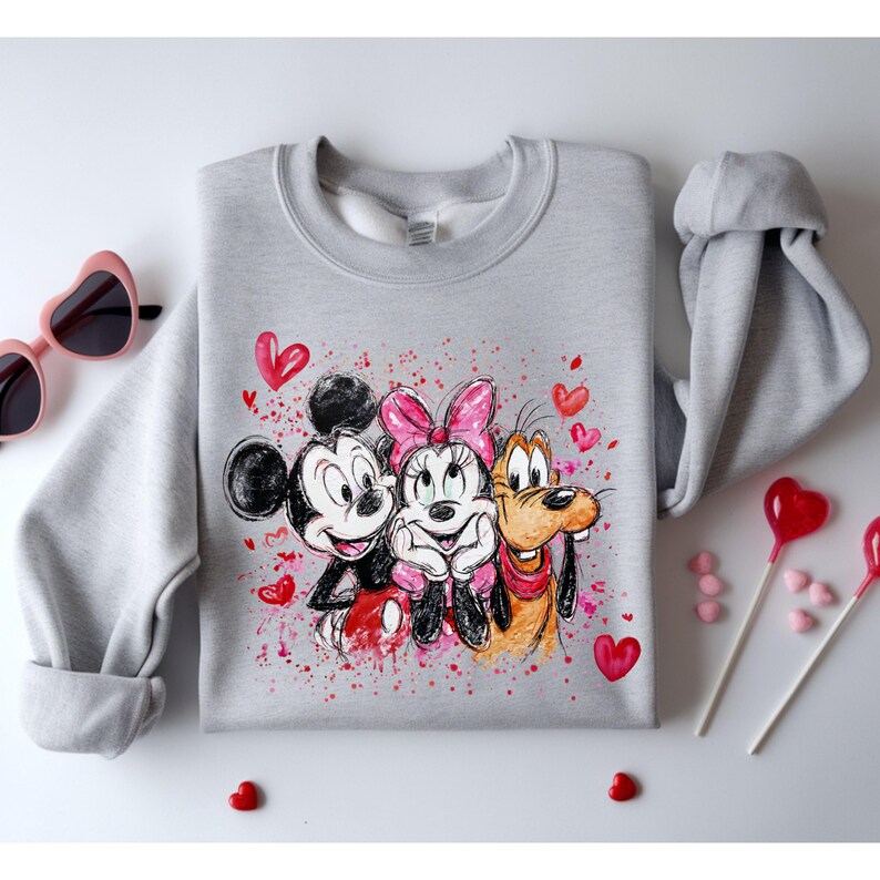 May include: A light gray sweatshirt featuring Mickey Mouse, Minnie Mouse, and Goofy, surrounded by red hearts and paint splatters. The cartoon characters are smiling and hugging. Pink heart-shaped sunglasses and heart-shaped lollipops are in the background.