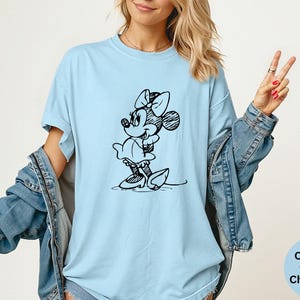 May include: Light blue t-shirt featuring a black and white sketch of Minnie Mouse. The shirt has short sleeves and a relaxed fit. The text "Comfort Colors Chambray" is in a light blue circle in the bottom right corner.
