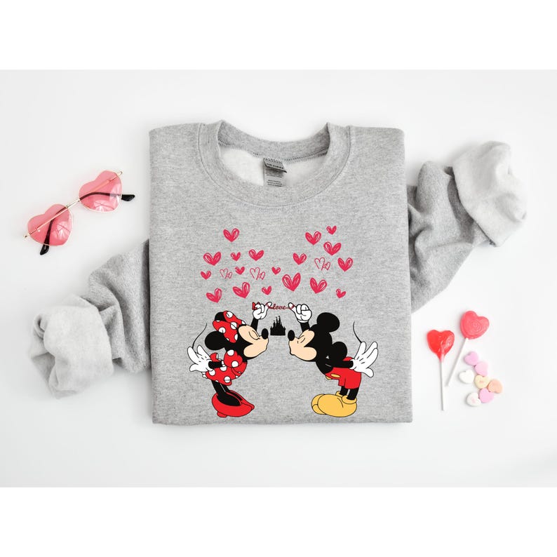 Puede incluir: Una sudadera gris con un gr&aacute;fico de Mickey y Minnie Mouse bes&aacute;ndose, rodeados de corazones rojos y la palabra "Love". El dise&ntilde;o incluye una silueta de castillo. Tambi&eacute;n se ven gafas de sol rosas en forma de coraz&oacute;n y piruletas.