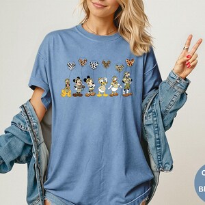 K&ouml;nnte beinhalten: Blaues Jeans-T-Shirt mit einer Grafik von Zeichentrickfiguren, die gemusterte Ballons halten. Zu den Figuren geh&ouml;ren ein Hund, Mickey Mouse, Minnie Mouse und Donald Duck. Das Shirt hat kurze &Auml;rmel und eine lockere Passform.