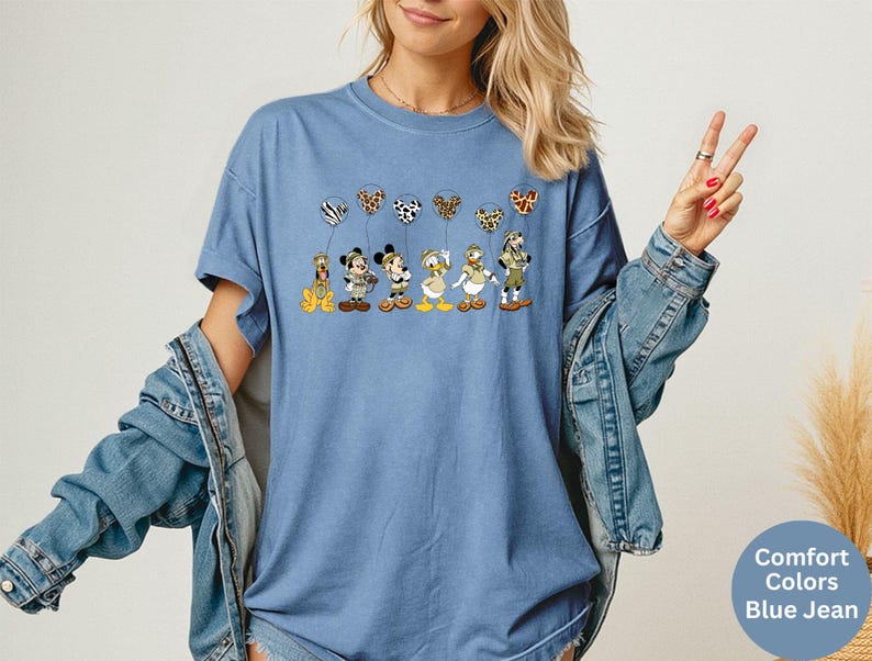 K&ouml;nnte beinhalten: Blaues Jeans-T-Shirt mit einer Grafik von Zeichentrickfiguren, die gemusterte Ballons halten. Zu den Figuren geh&ouml;ren ein Hund, Mickey Mouse, Minnie Mouse und Donald Duck. Das Shirt hat kurze &Auml;rmel und eine lockere Passform.