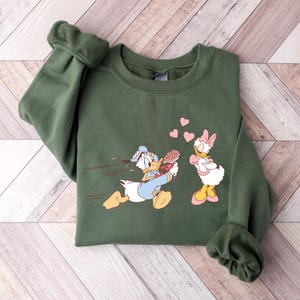 Op de afbeelding: Een groen sweatshirt met een cartoon afbeelding van Donald Duck die rent met een boeket bloemen naar Daisy Duck, die met gekruiste armen staat en een hartvormige wolk boven haar hoofd heeft. Er zweven roze harten in de lucht.