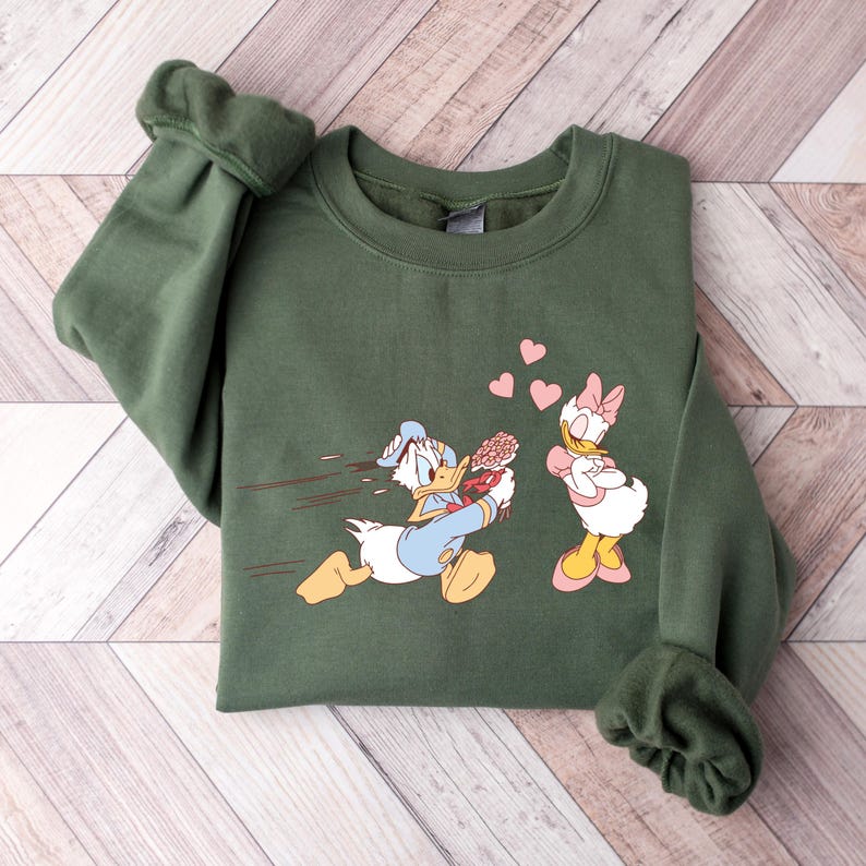 Op de afbeelding: Een groen sweatshirt met een cartoon afbeelding van Donald Duck die rent met een boeket bloemen naar Daisy Duck, die met gekruiste armen staat en een hartvormige wolk boven haar hoofd heeft. Er zweven roze harten in de lucht.