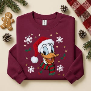 May include: Burgundy crewneck sweatshirt featuring an embroidered Donald Duck wearing a Santa hat and a green and red striped scarf. The design includes snowflakes, holly, and gold sequins. A gingerbread man and pinecones are visible.