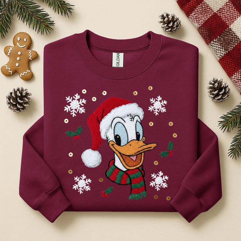 May include: Burgundy crewneck sweatshirt featuring an embroidered Donald Duck wearing a Santa hat and a green and red striped scarf. The design includes snowflakes, holly, and gold sequins. A gingerbread man and pinecones are visible.