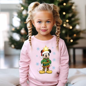 Pode incluir: Uma camisola rosa com um design do Mickey Mouse com tema natalino. Mickey est&aacute; usando um gorro de Papai Noel, um su&eacute;ter verde e enfeites dourados. O design inclui flocos de neve e uma estrela na cabe&ccedil;a do Mickey. Uma &aacute;rvore de Natal est&aacute; no fundo.