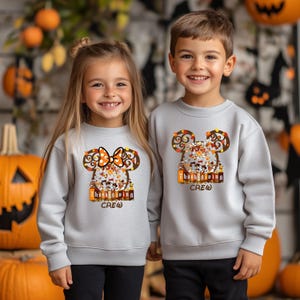 Puede incluir: Dos ni&ntilde;os con sudaderas grises claras con un dise&ntilde;o tem&aacute;tico de Acci&oacute;n de Gracias. El dise&ntilde;o presenta una cabeza con forma de Mickey Mouse con hojas de oto&ntilde;o y las palabras "Thanksgiving Crew". La sudadera de la ni&ntilde;a tiene un lazo naranja.