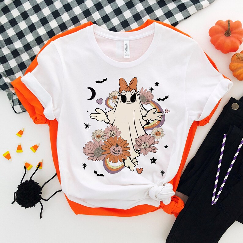May include: White t-shirt with a cartoon ghost wearing a pink bow and surrounded by flowers and stars. The ghost is white with a black outline and has a surprised expression. The flowers are pink, orange, and yellow. The stars are black.